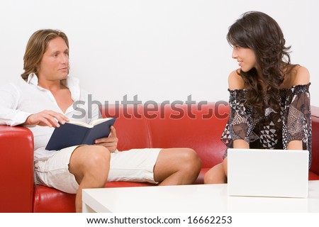 Young couple sitting on the sofa at home, he reads a book, she works  with the laptop