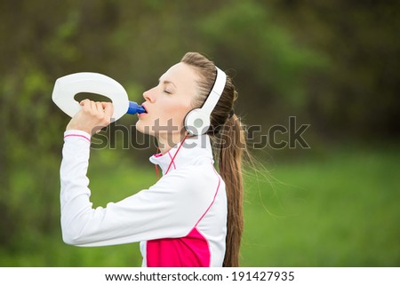 Sporty woman drinking water after working out, jogging on a hot day over green nature background