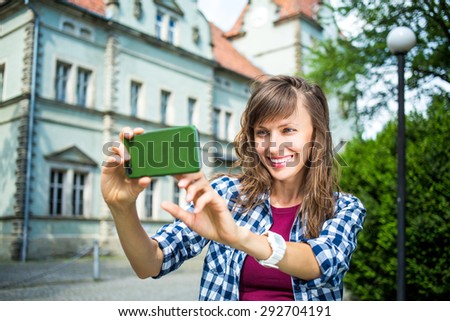 Smiling young woman taking selfie picture with smart phone camera outdoors