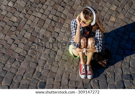 Top view young woman sitting on paving stones enjoying the sunshine and listening music, tourist girl in bright glasses relaxing outdoors, full length, copy space
