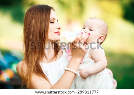 Beautiful young mother is feeding her baby from a bottle outdoor. Outdoor Portrait of happy family