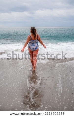 A fat woman in a bathing suit enters the water during the surf. Alone on the beach, Gray sky in the clouds, swimming in winter. Photo stock © 