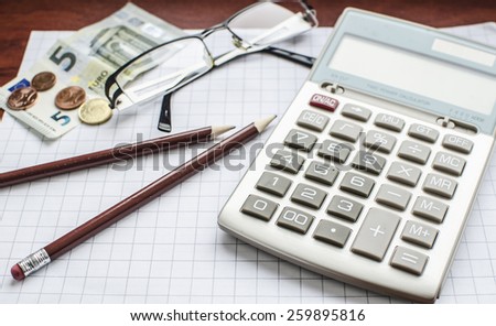 A calculator laying on the office paper with pencils, euro banknotes and coins and transparent glasses