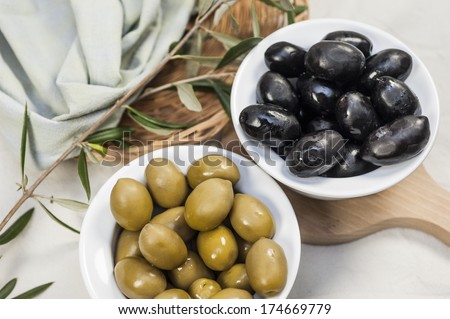 black and green olives are in separate white bowls next to an olive tree twig