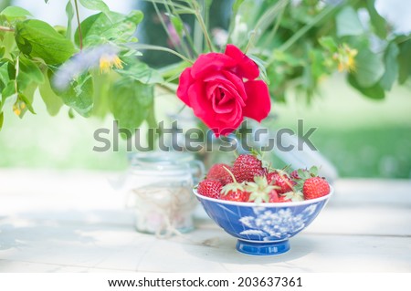 A pot of strawberries and colorful sugars under the shadow of a vase full of flowers, in a shabby chic mood.