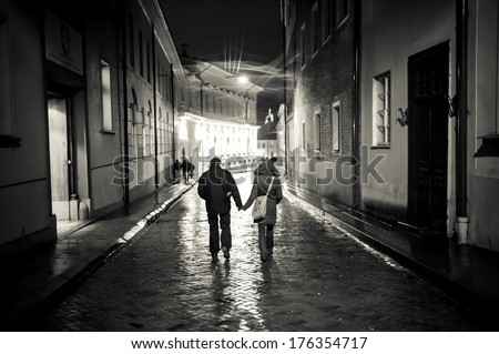 A young couple holding hands walking at night in old town street, cobbled street wet from rain, autumn evening, black and white. Visible only silhouettes of models, there is no need model release.
