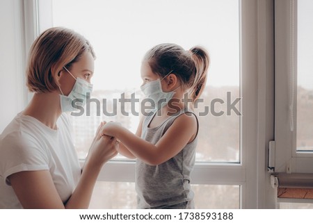 Family Mom and Daughter in Medical Mask. Young Woman and Child little girl sitting by the Window in Protective Masks against the Virus. Mom carefully corrects the mask on the face of the child        