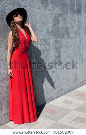 Beautiful girl in a red dress and hat with glasses . Beautiful brown-haired woman in a high hat in red dress and black sunglasses. A woman in a dress on a background of gray stone wall.