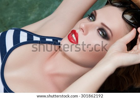 Beautiful girl near the water lifted glasses.The concept of beauty make-up. Close portrait of a woman in a striped bathing suit, her eyes looking straight at the camera. Beautiful makeup ,  red lips.