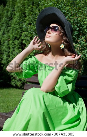 Beautiful model with long hair in the park holds hands a hat. The girl in the green dress, black hat and sunglasses