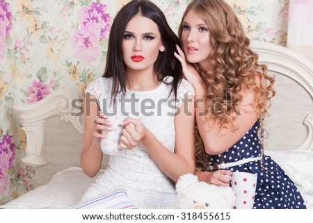 Two pretty girl-friends talk and drink tea .Happy girls drinking tea and gossiping in home. Two girlfriends blonde and brunette drinking tea in bed and talking. concept female friendship.
