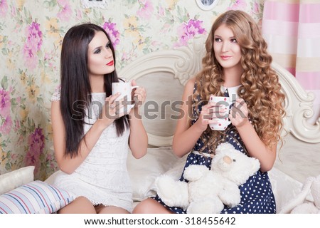 Two pretty girl-friends talk and drink tea .Happy girls drinking tea and gossiping in home. Two girlfriends blonde and brunette drinking tea in bed and talking. Comfort in the house, friends