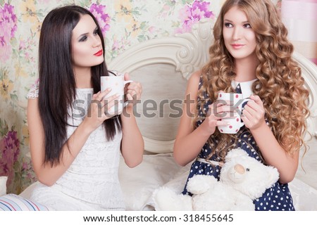 Two pretty girl-friends talk and drink tea .Happy girls drinking tea and gossiping in home. Two girlfriends blonde and brunette drinking tea in bed and talking. Girls and boys, teenagers discuss