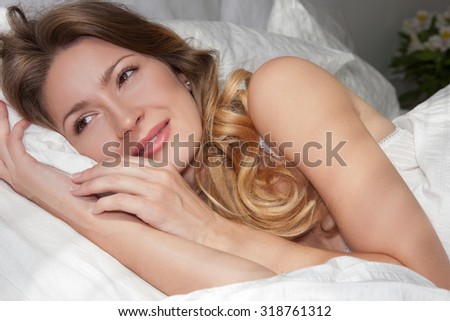 Close portrait of a beautiful girl lying in her bed, her arms wrapped around the pillow, her eyes slightly open, her face gentle smile. Bed and woman illuminated by the sun through the blinds
