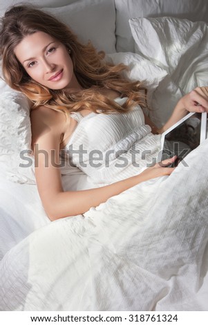 Woman in bed in the morning with a tablet in her hands, white bed bright bedroom, curly hair on her head.Beautiful girl looks  the camera, the blonde with a bed, a gentle make-up on her face