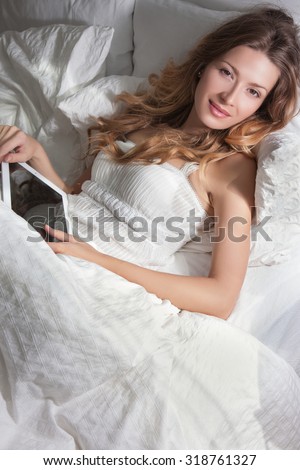 Attractive woman in bright pajamas lying in bed reading an e-book, morning. Warm and comfortable bed, a beautiful blond woman lying in bed, brown eyes and curly hair on her face. Concept Morning.