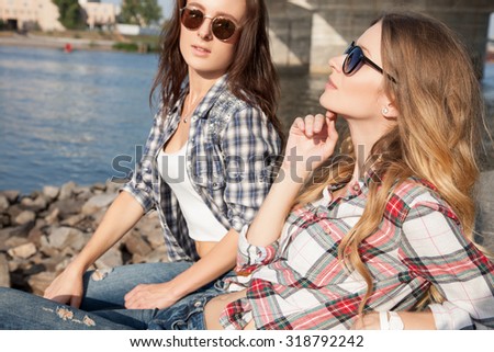 Sporting an attractive woman relaxing by the river near the bridge,the sun is shining in  faces,they bask in the sun.Two friends sitting on the promenade near the pier and looking into the distance