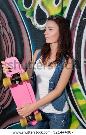 The girls on skateboards. Slim and beautiful woman holding a pink skate. Attractive girl in jeans and shirt holds the skate. Stylish girl goes in for sports and fun riding on a skateboard