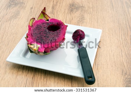 Half a dragon fruit cut open to show the red flesh. A ball of flesh has been removed. All on a white plate displayed on a wooden background.