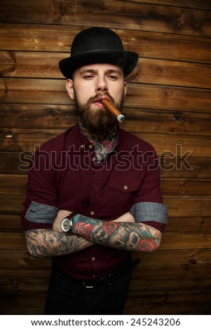 Brutal male with tattoo standing in front if wooden wall and smokes vigar