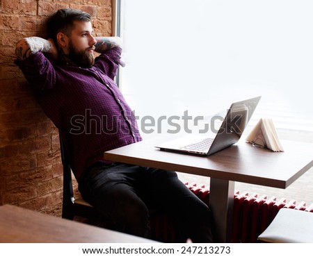 Fashionable male with tattooes sitting at the table in coffe shop with a laptop
