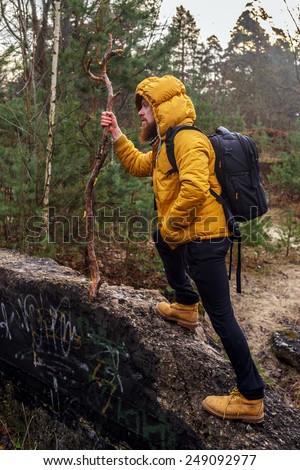 Man with beard  in yellow jacket, yellow boots and black pants with bacpack and stick  walking in a forest.