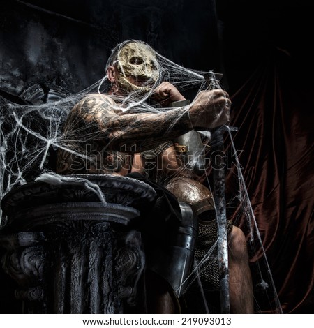 Muscular man with skull on his face and sword in his hand posing on the throne in spiders web on gray background.