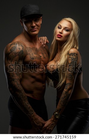 Tattooed guy with naked torso posing with blond tattoed female. Isolated on grey