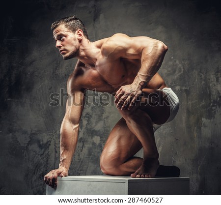 Bodybuilder guy posing on white podium on his knees. Isolated on grey background.