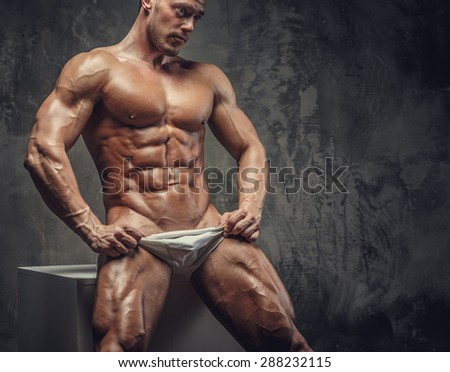 Awesome shirtles muscular guy sitting on white podium. Isolated on grey background.