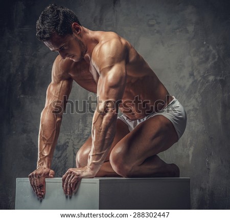 Muscular guy in white panties standing on his knees on white podium in studio.