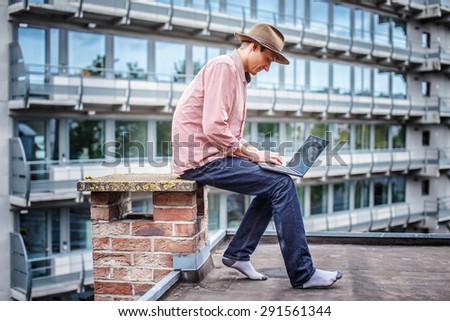 Man in blue jeans and pink shirt sitting on the roof with laptop.