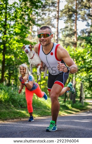 Smiling sportsman in sunglasses on the run with small dog in his hand. Running woman on background.