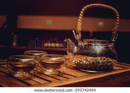Glass chinese teapot with herbs and tea cups on wood tray.