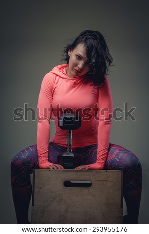 Fitness woman with short black hair in colorful sportswear sitting on wooden box with dumbell.