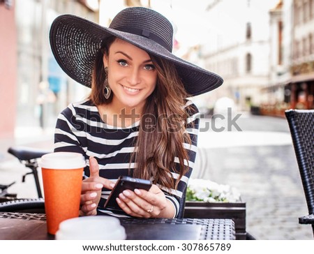 Smiling young woman in black shirt with white stripes and big black summer hat sitting at the table in summer cafe.