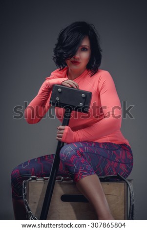 Young fitness woman with black hair in pink shirt and violet pants sitting on wooden box and holding big black hummer.