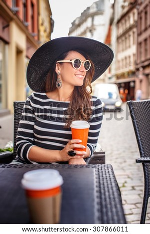 Beautiful young lady in big summer hat and in dress with stripes drinking coffe outside in summer cafe