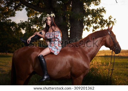 Fashion brunette woman in colorful dress and long leather boots sitting on brown horse over nature scene on background.