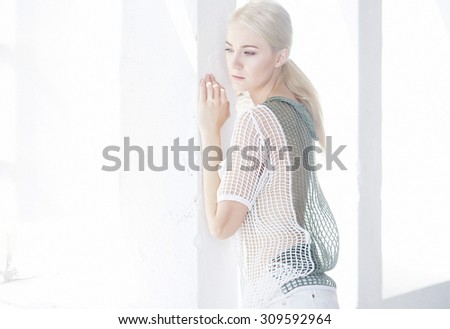 Sexy blond woman in white clothing posing near the wall with lights from window.