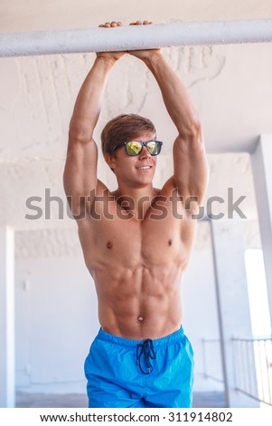 Smiling young shirtless guy in sunglasses posing with hands up.