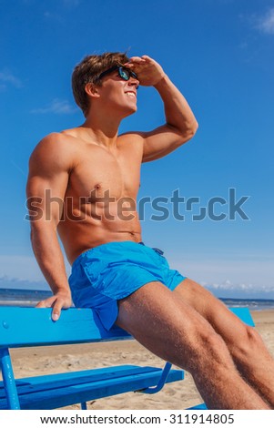 Awesome muscular young guy in blue swim shorts sitting on bench over blue sky and sea.