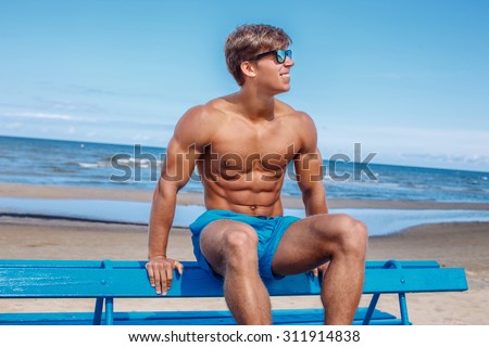 Awesome muscular young guy in blue swim shorts sitting on bench over blue sky and sea.