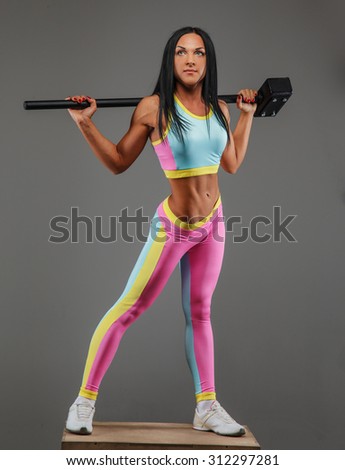 Slim brunette female in colorful sportswear posing over grey background with hummer on her shoulders.