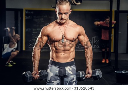 Muscular guy in military pants doing exercises with dumbells in a gym.