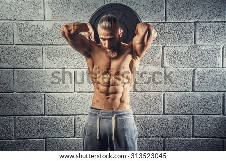 Shirtless muscular man with beard holding disk weight behind his head. Grey wall from bricks background.