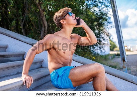 Muscular man in blue swimming shorts talking by smartphone.