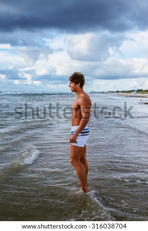 Muscular guy in swimming shorts standing in water on a beach over cloudy sky.