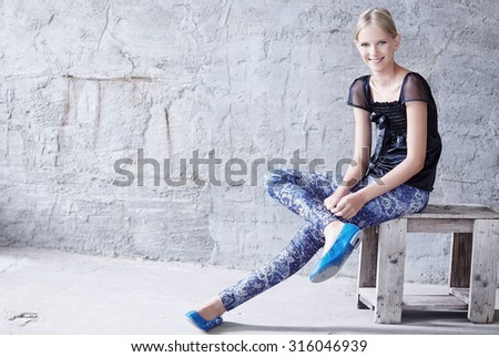 Young blond woman in casual clothing sitting on wooden chair over grey wall.
