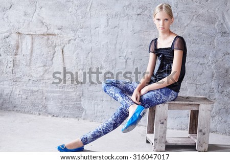 Young blond woman in casual clothing sitting on wooden chair over grey wall.